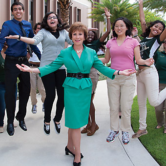 USF System President Judy Genshaft stands in front of students, who are jumping in the background.