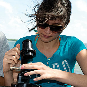 A student adjusts a microscope while outside by the water.