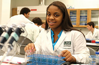 Student in the STEM Academy poses in a white lab coat in front of test tubes.