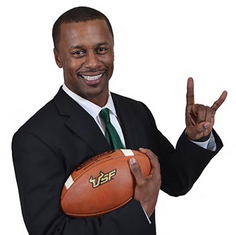 Willie Taggart holding a football while making the Bulls Horns sign with his hand.