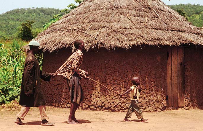 A young child holding a long stick leads two blind men.