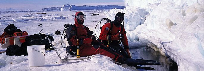 Two divers sit on the edge of a hole in the ice.